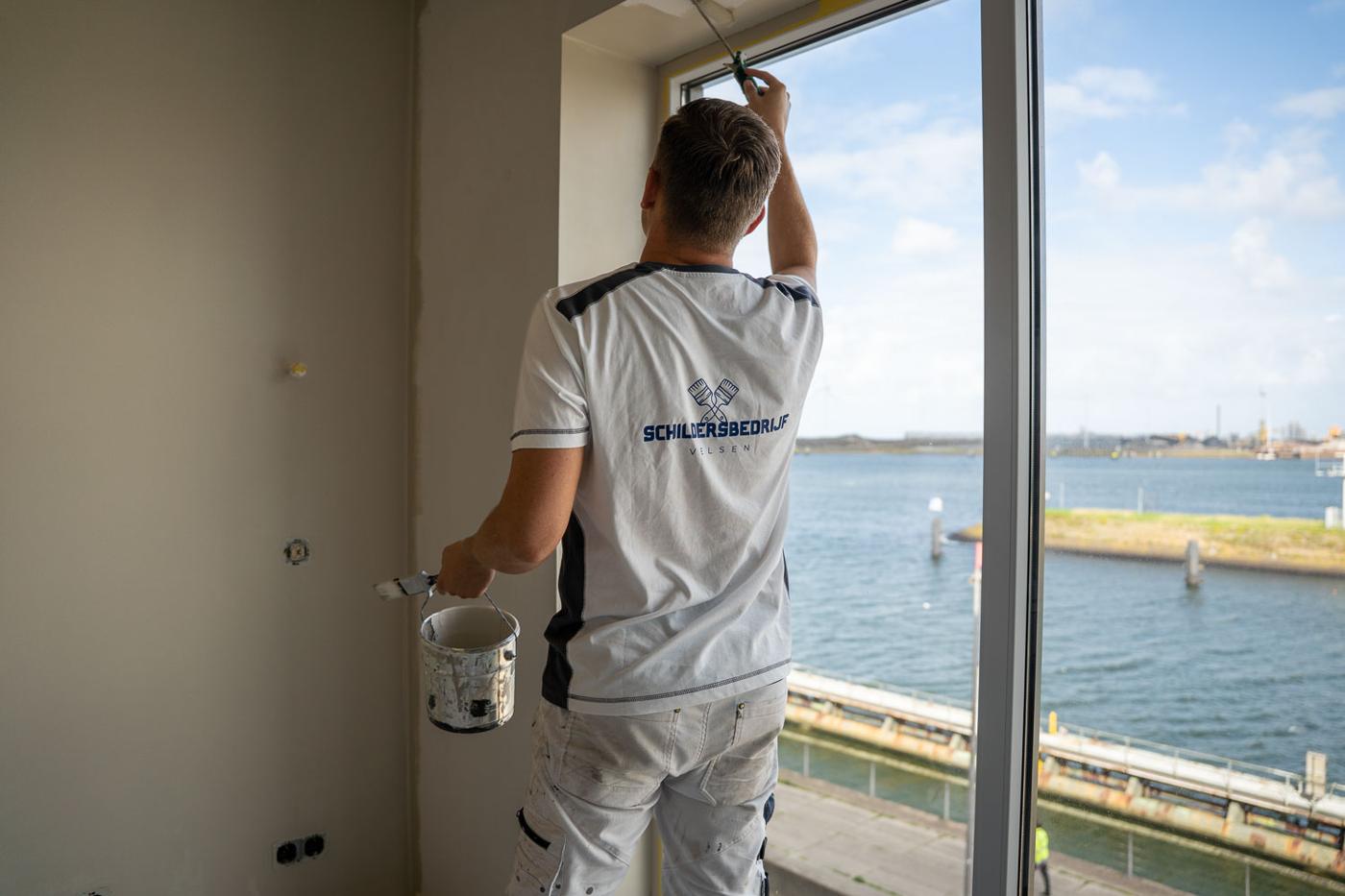 Schilder lakt een kozijn bij het raam, haven van IJmuiden op de achtergrond